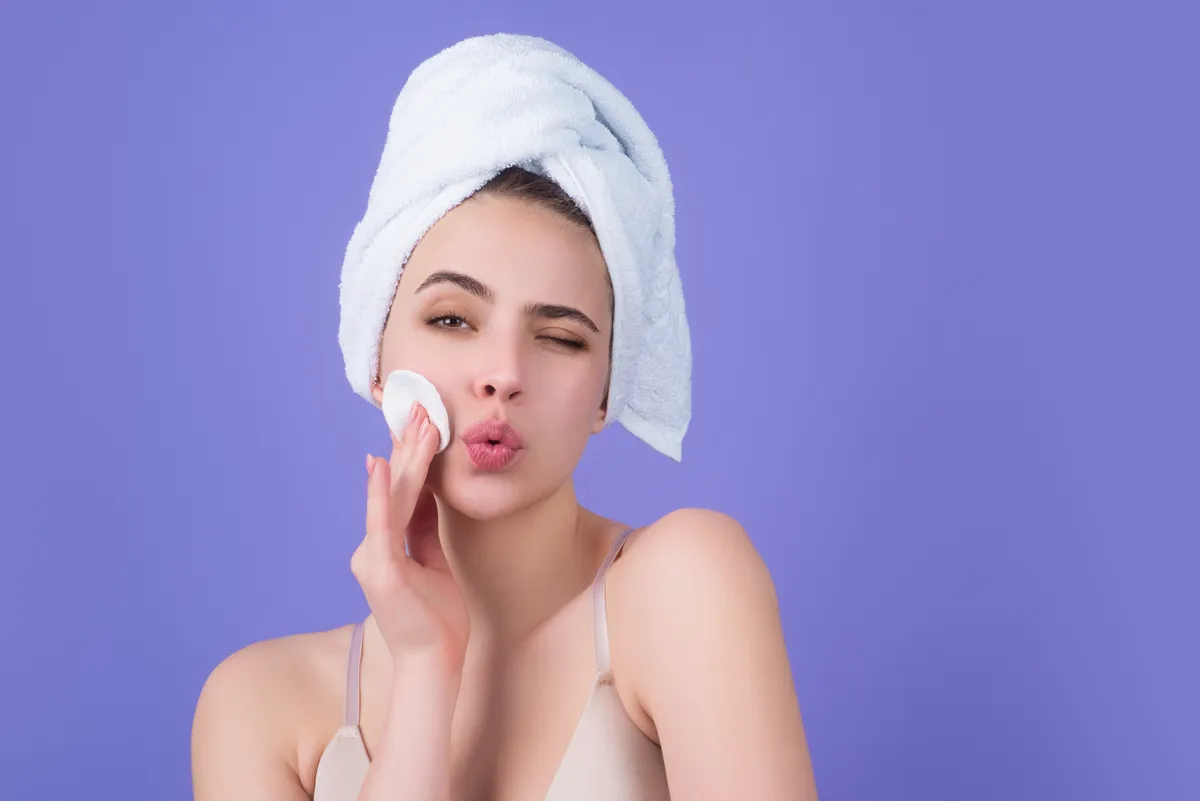 bőrfeszesítő trükk, fiatal nő, bőrápolás, sminklemosó vatta, spa, szépségápolás, lila háttér