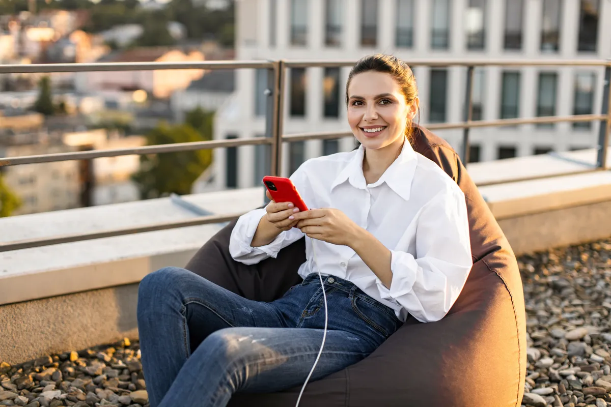 Nő mosolyogva ül egy babzsákfotelben miközben tölti a telefonját