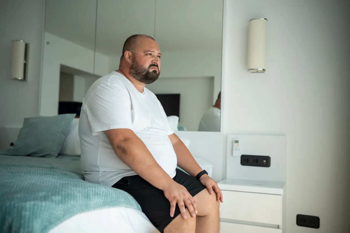 Kövér férfi ül az ágyon a hálószobában