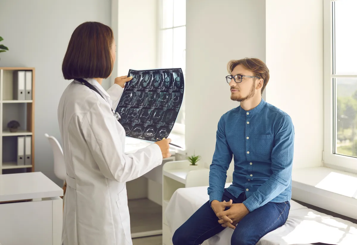 Fiatal férfi térdfájdalom miatt ül egy orvosi rendelőben, ahol a doktornő a röntgenfelvételeit tartja a kezében