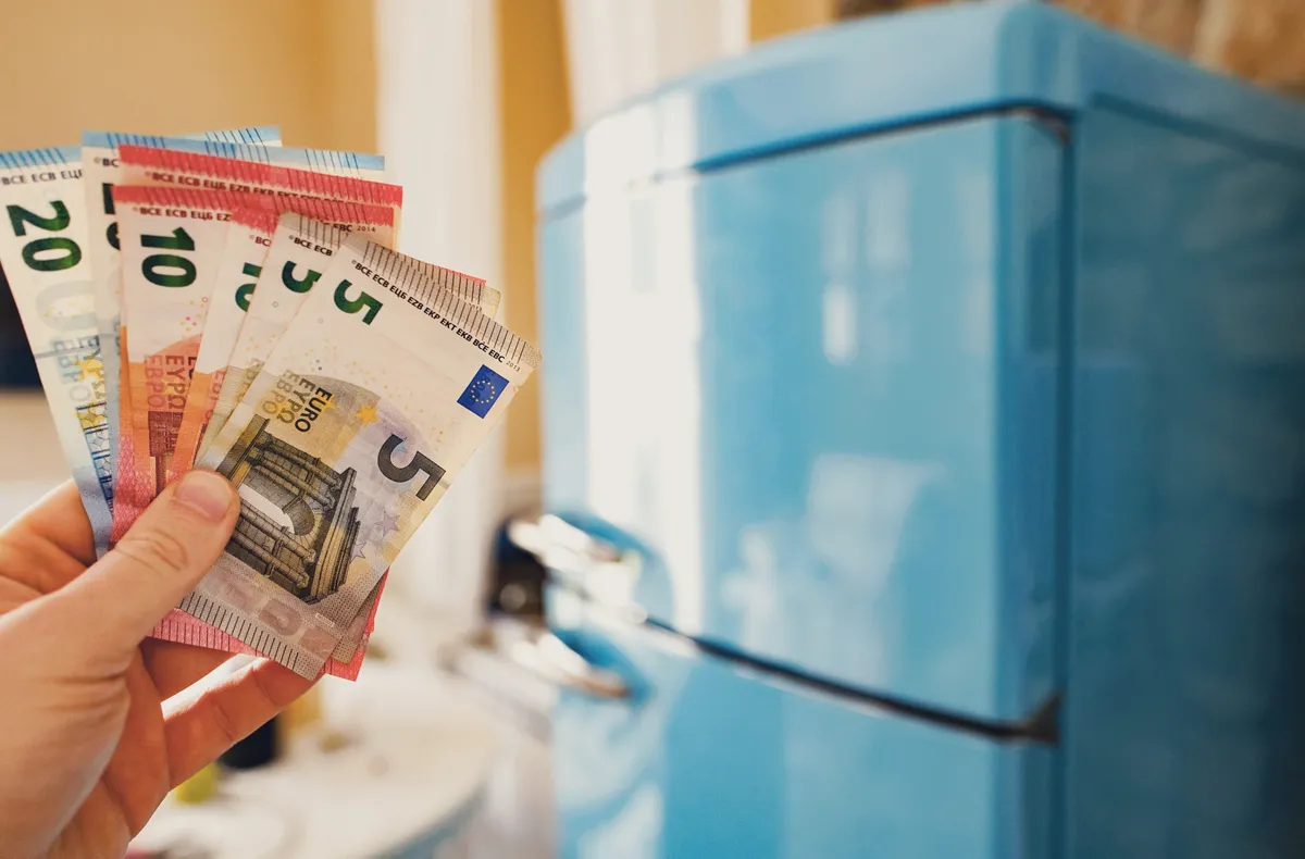Kék hűtőszekrény előtt pénzt tartó kéz, amely a hűtőszekrény fogyasztása miatti kiadások csökkentését és a helyes használattal való spórolást jelzi.