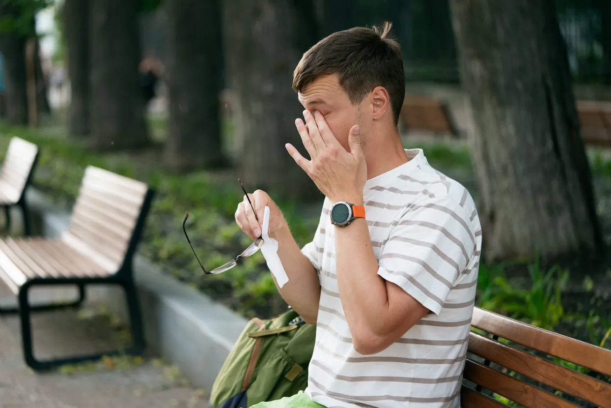 Szemét vakaró szemüveges fiatal férfi ül egy parki padon