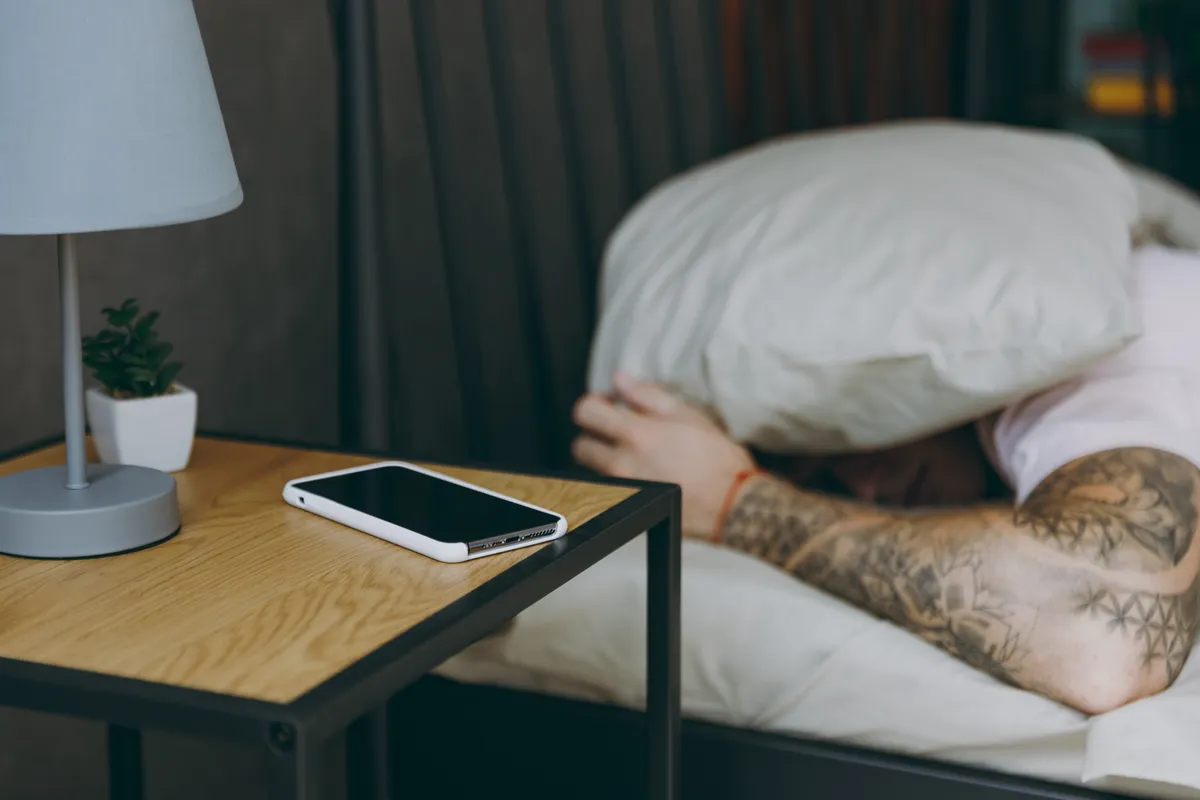 jetlag, tetovált férfi alszik, párnával a fején, előtérben telefon