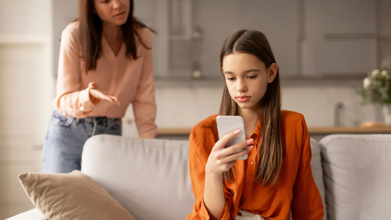 Digitális házirend szabályai szerint okostelefont használó kislány a kanapén mögötte a figyelő anyjával