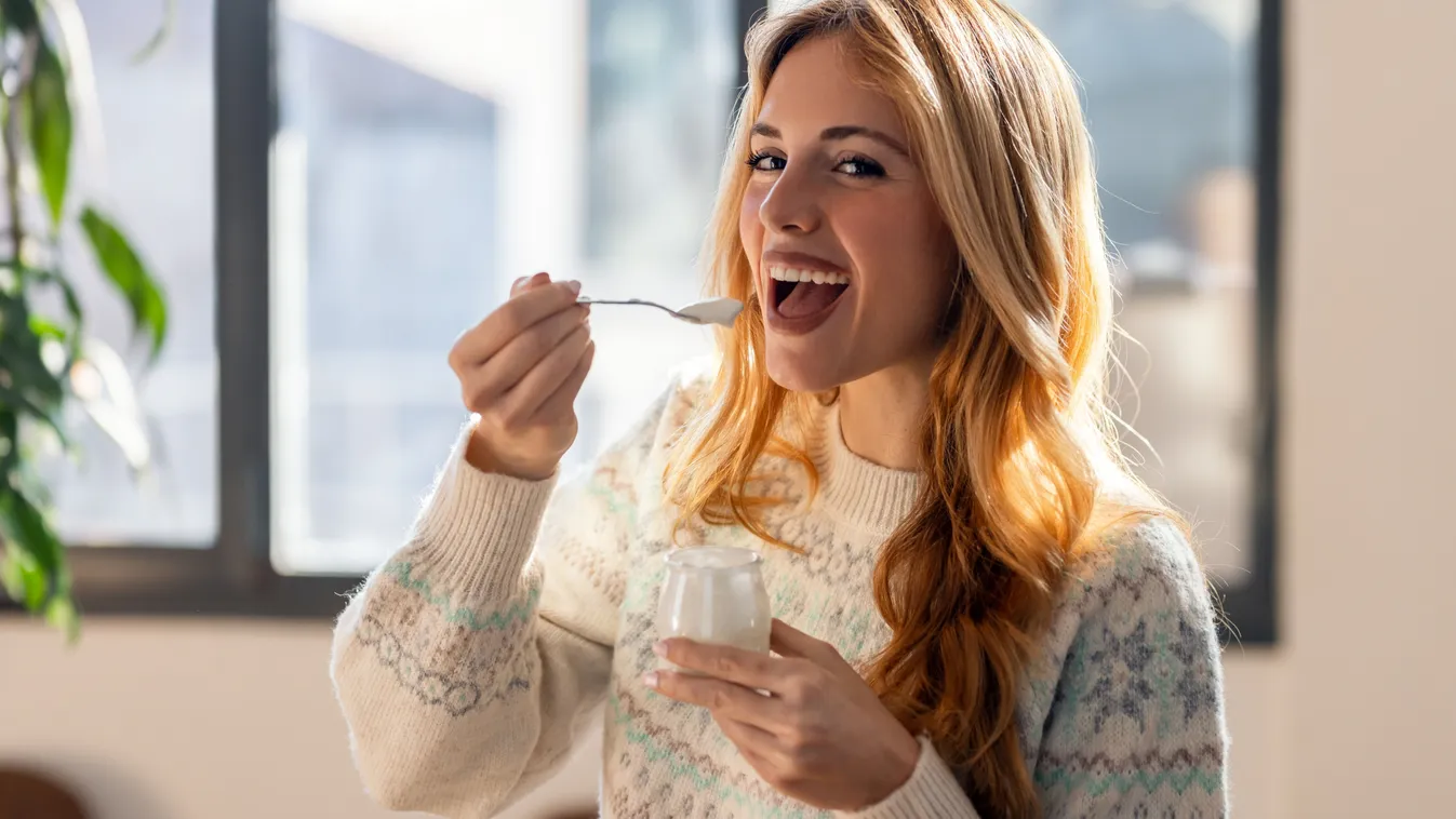 Fiatal szőke nő vidáman kanalaz egy üveg joghurtot