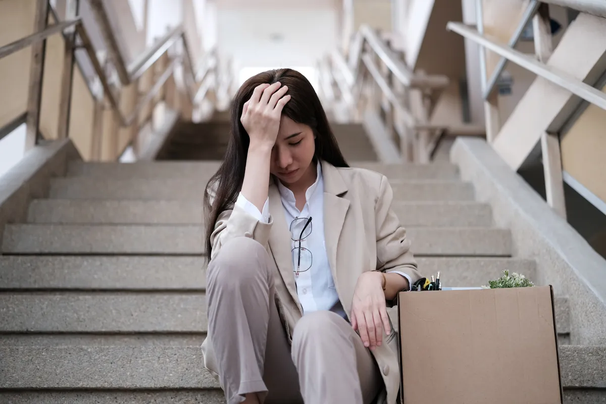 A kirúgása után egy nő elkeseredetten ül a lépcsőn, mellette egy doboz tele a holmijaival.