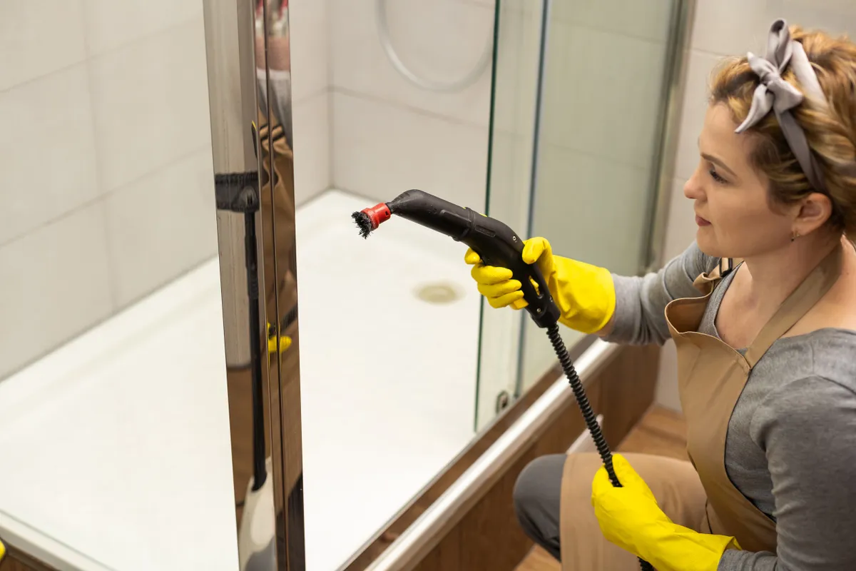 Fürdőszoba-takarítás: egy hölgy gőzzel zuhanykabint tisztít