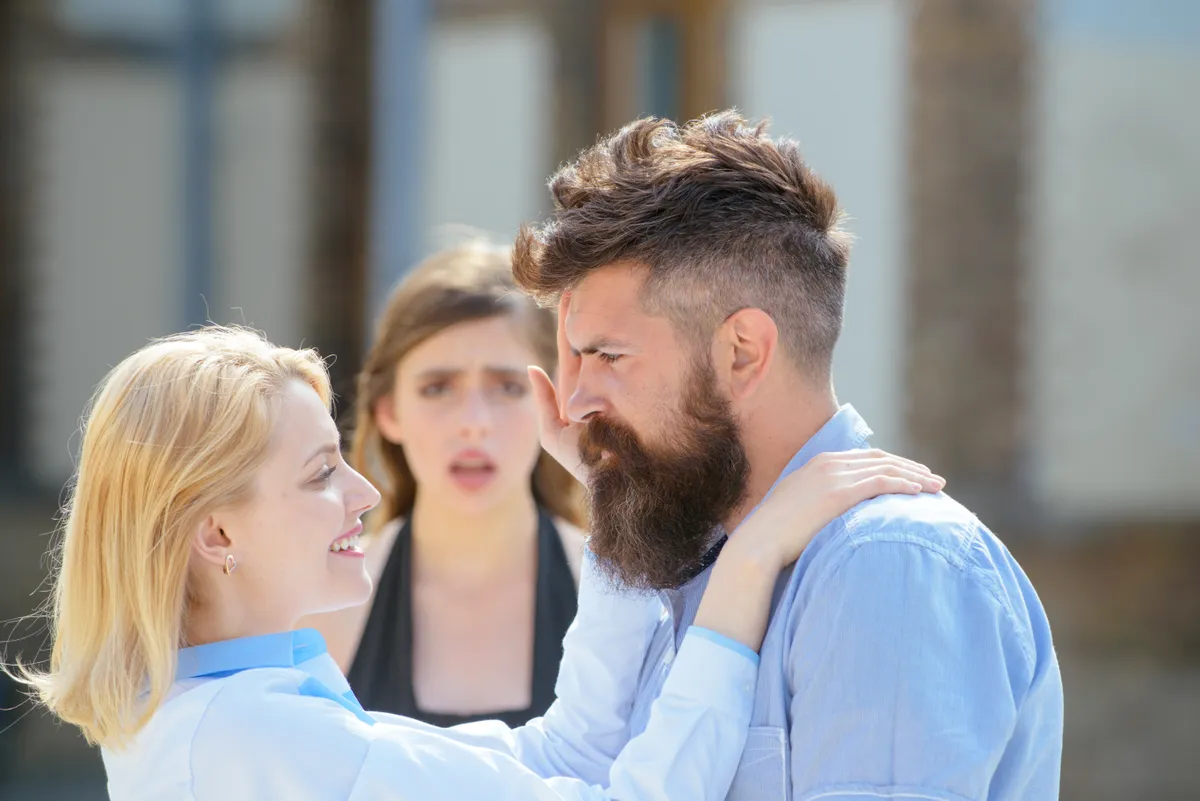 Hűtlenség miatt feldúlt nő nézi a párját és egy szőke nőt.