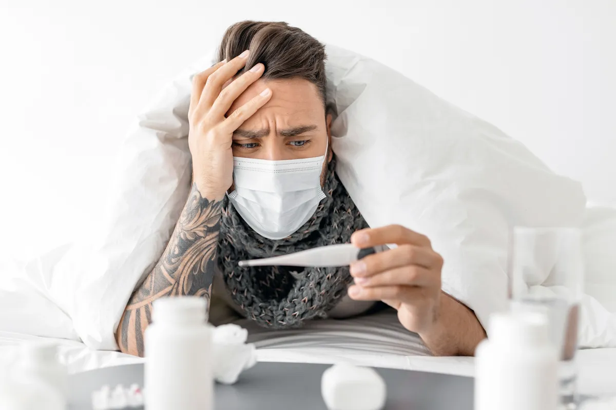 Influenza, Covid és megfázás hasonló tüneteit produkáló, lázat mérő beteg férfi sálban és maszkban a takaró alatt