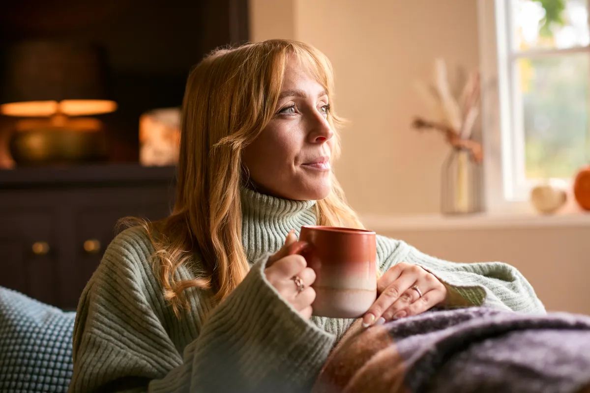 Nő ül a kanapén karácsonyi fényekkel megvilágított lakásban, kezében egy bögre.