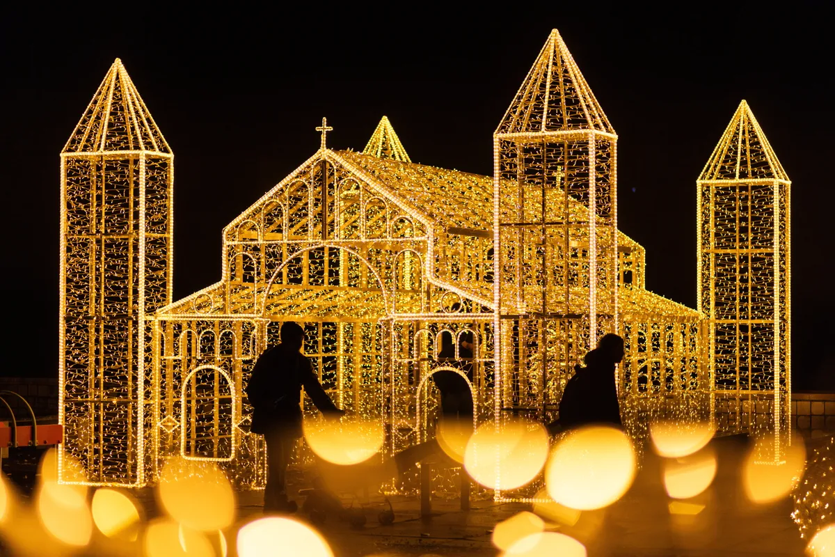 két ember áthaladó sziluettje egy nagy méretű templomot ábrázoló fényinstalláció előtt.