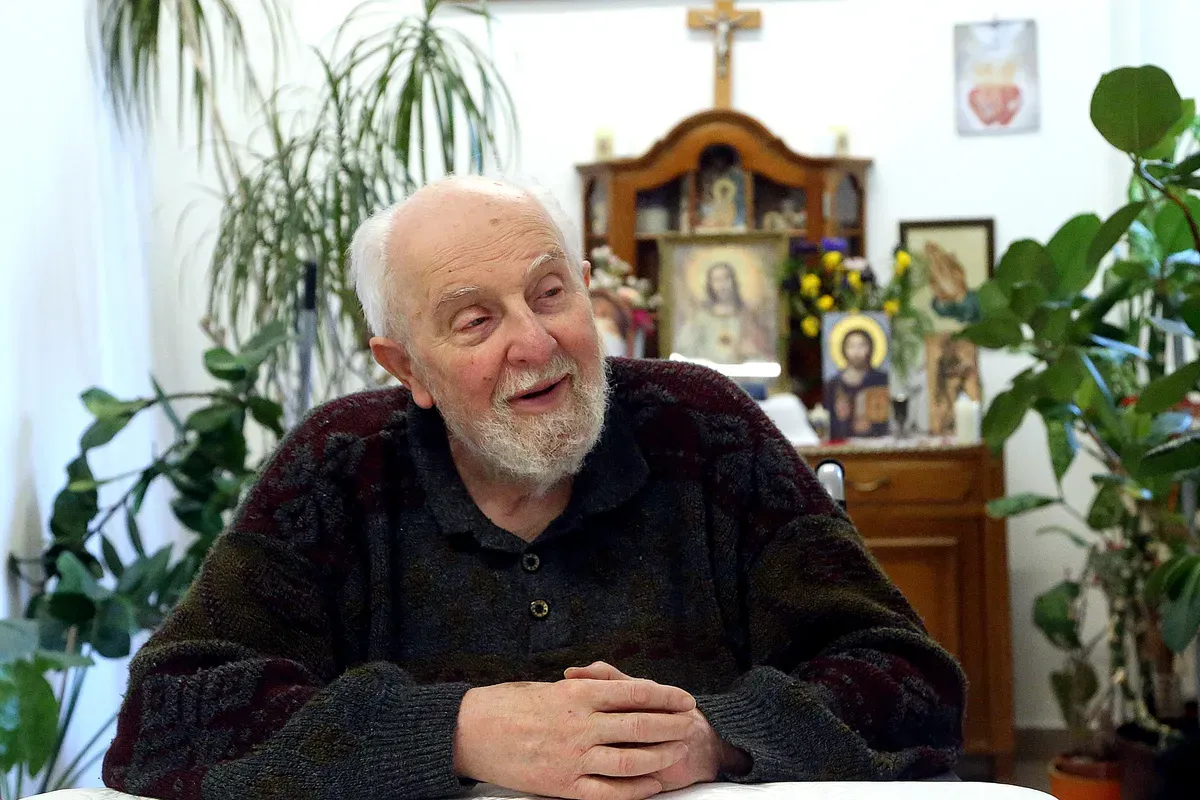 Kardos atya egy asztalnál ül, fekete pulcsiban, mögötte egy oltár kereszttel.