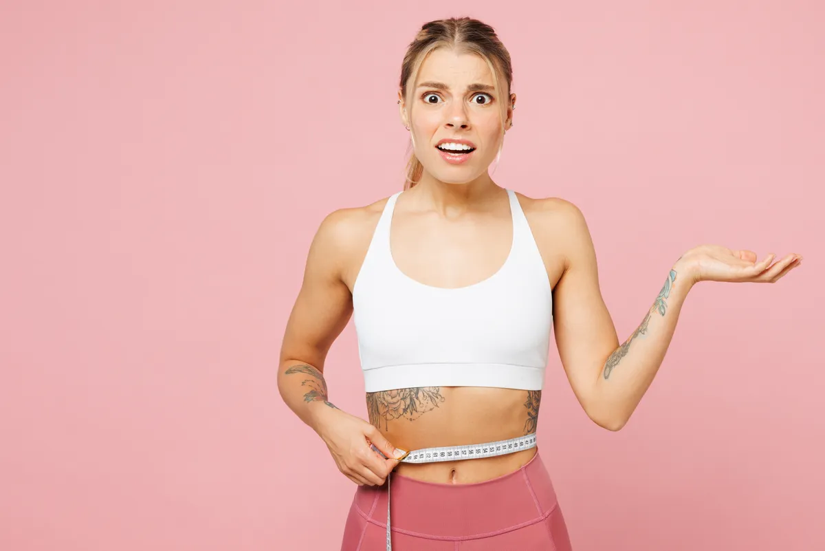 Fiatal nő meglepődve néz a kamerába derekán fehér mérőszalaggal