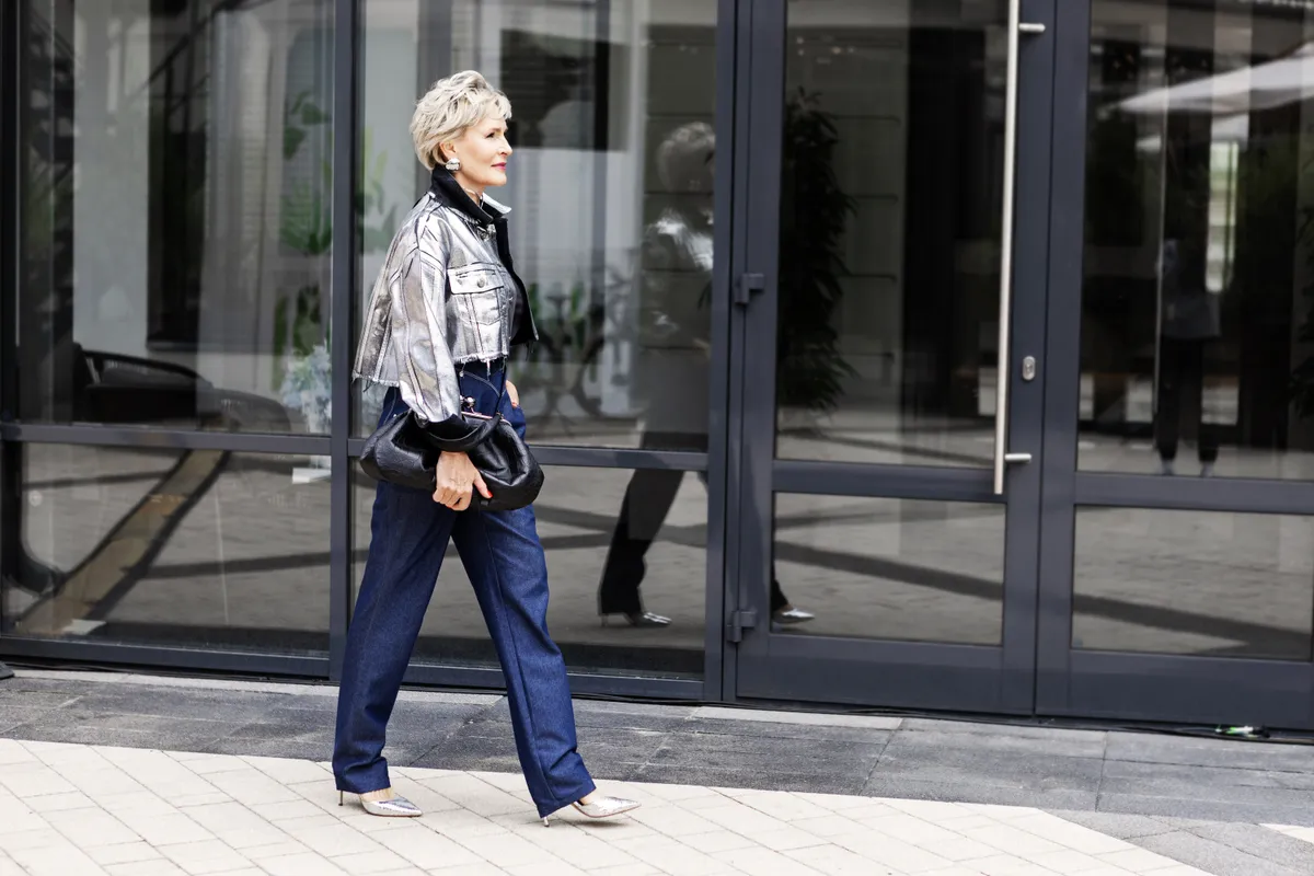 Középkorú stílusos nő bő szárú nadrágban és rövid ezüst kabátban sétál az utcán.