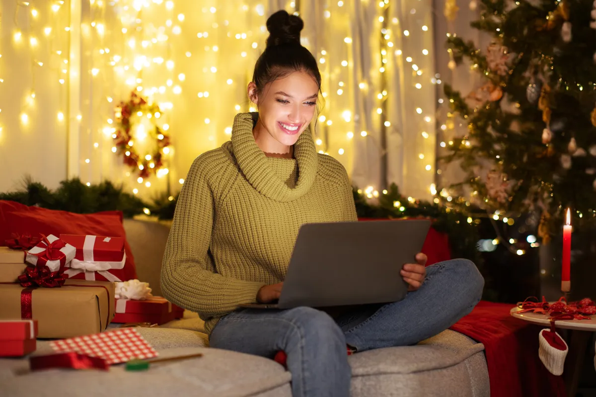 Fiatal nő ül a kanapén, mögötte fényfüzérek világítanak. Mosolyog, mert valószínűleg tud spórolni a karácsonyi fényekkel.