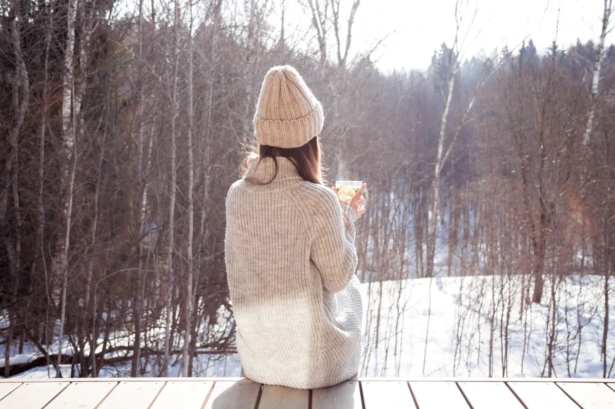 A téli divatnak megfelelően kiegészítőket viselő nő ül és nézi a havas tájat.