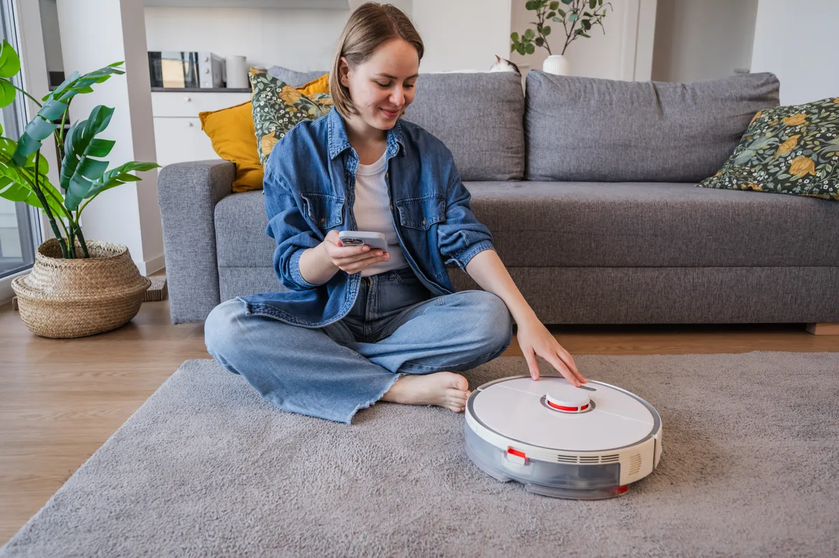 Egy fiatal nő épp egy robotporszívót indít be a szőnyegen.