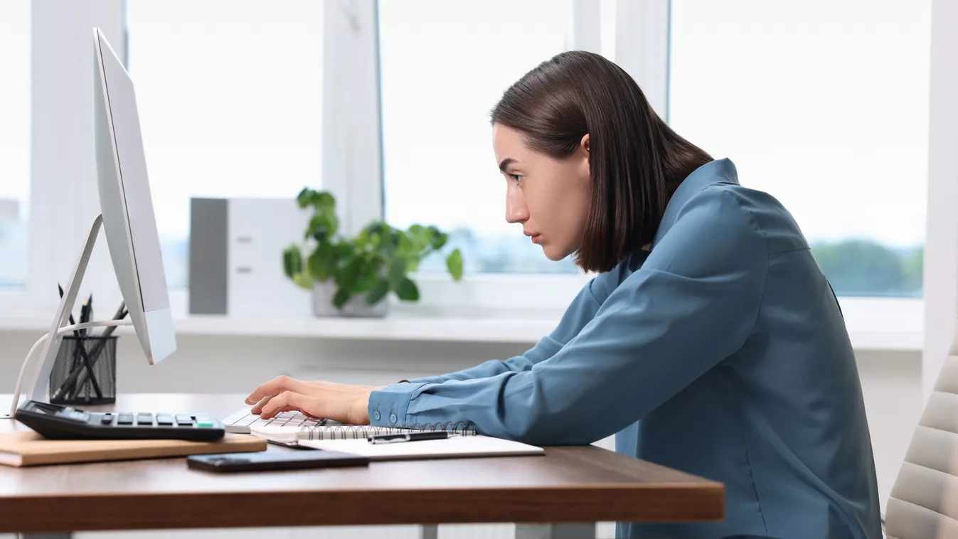 Banyapúpos fiatal nő görnyed az irodában a számítógép előtt