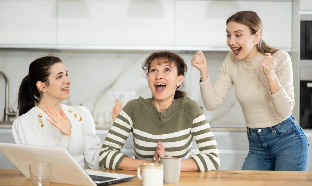 Három nő örül és hálálkodik egy konyhaasztalnál egy laptop előtt, mert a vonzás törvénye meghozta a szerencséjüket. 