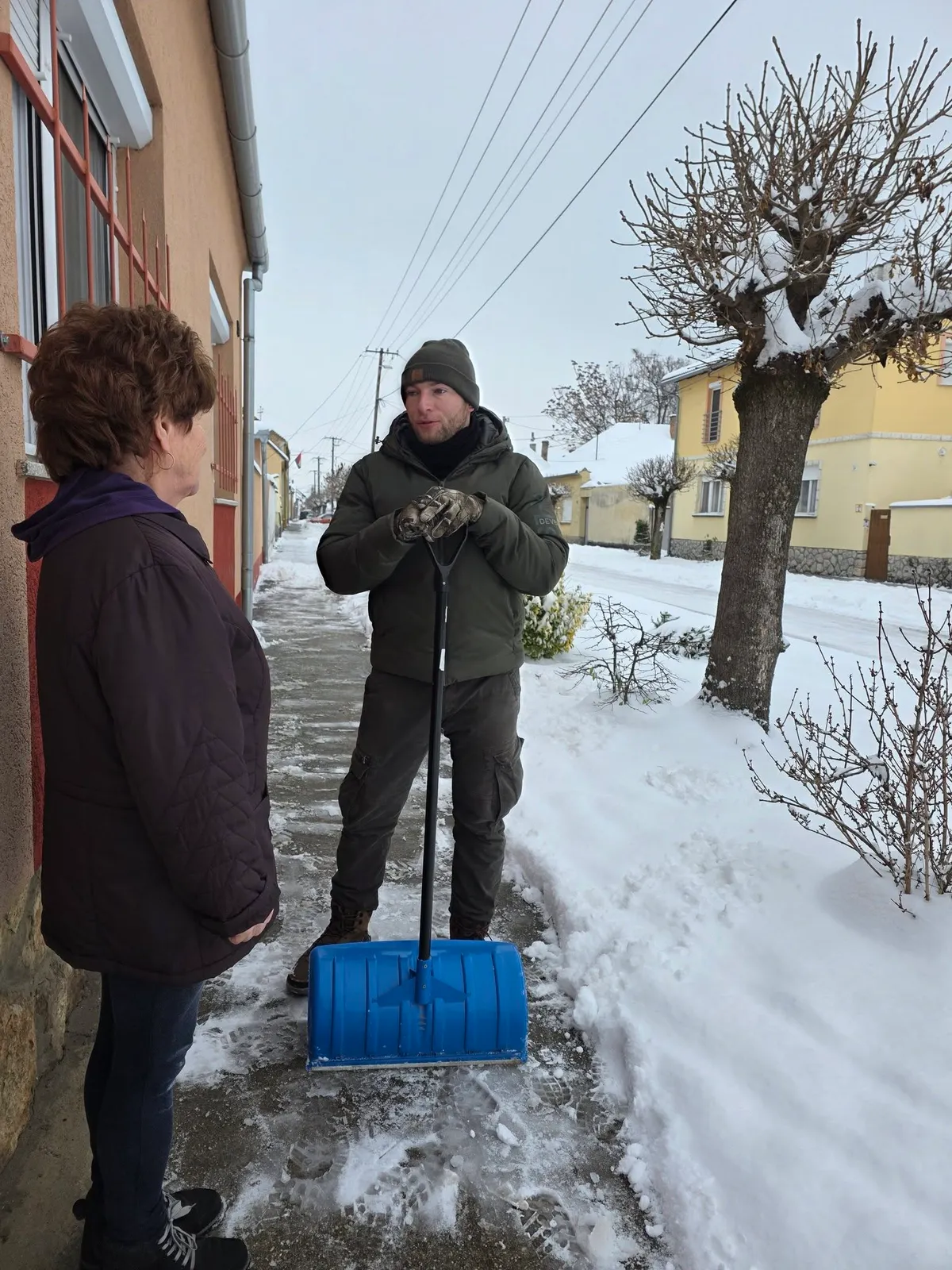Nyugdíjas asszony áll az utcán, ahol mindent elfed a hó. Mellette egy férfi támasztja a hólapátot és beszél. 