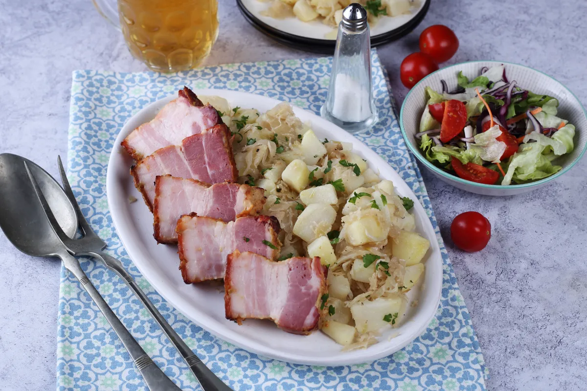 Lassan sült mézes-mustáros császárszalonna káposztás burgonyával egy tányéron, ami a legfinomabb szalonnás ételek közé tartozik. 