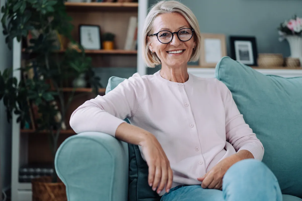 50 feletti szemüveges nő elegáns frizurával ül a kanapén.