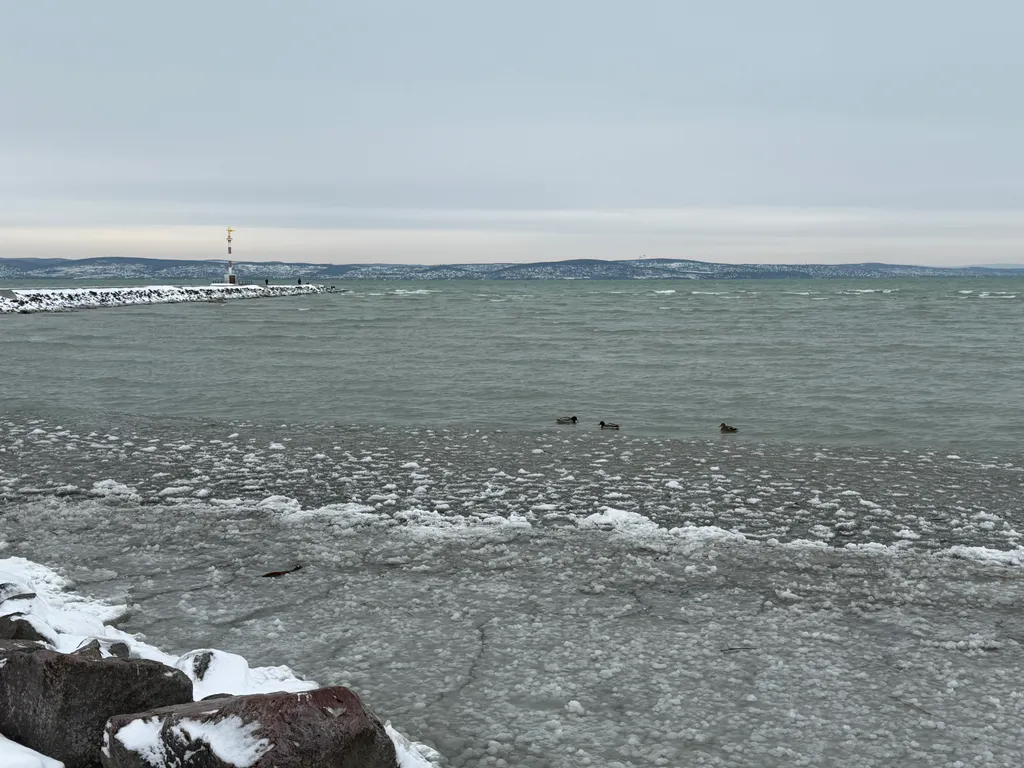 A képen a jeges Balaton látható, a távolban pedig a A Béke jóságos angyala szobor emelkedik a magasba.