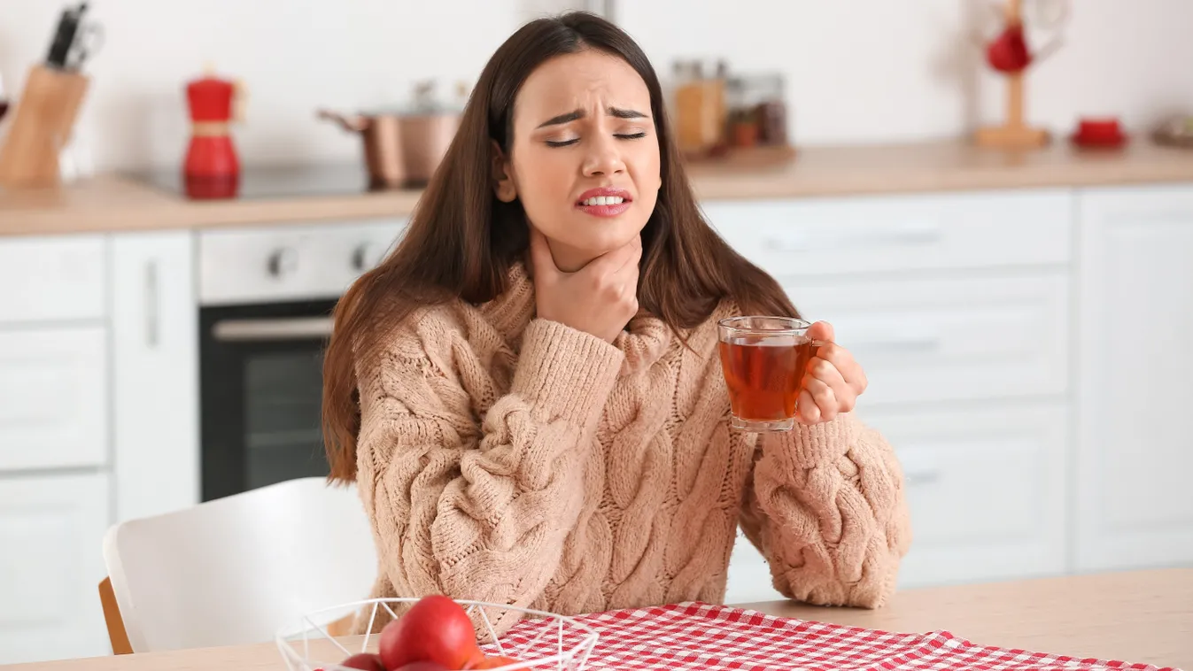 Torokfájással küzdő, meleg teát fogyasztó fiatal nő a konyhai étkezőasztalnál