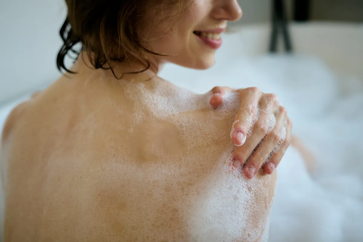 Fiatal nő forró zuhanyt vesz, közben habos a válla és a bőre.