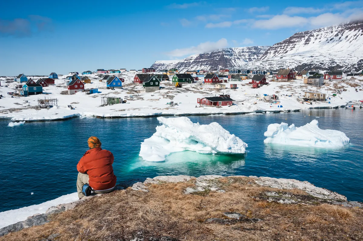 Piros dzsekis turista Qeqertarsuaq színes házait, és a borjadzó jégtömböket csodálja Grönlandon.