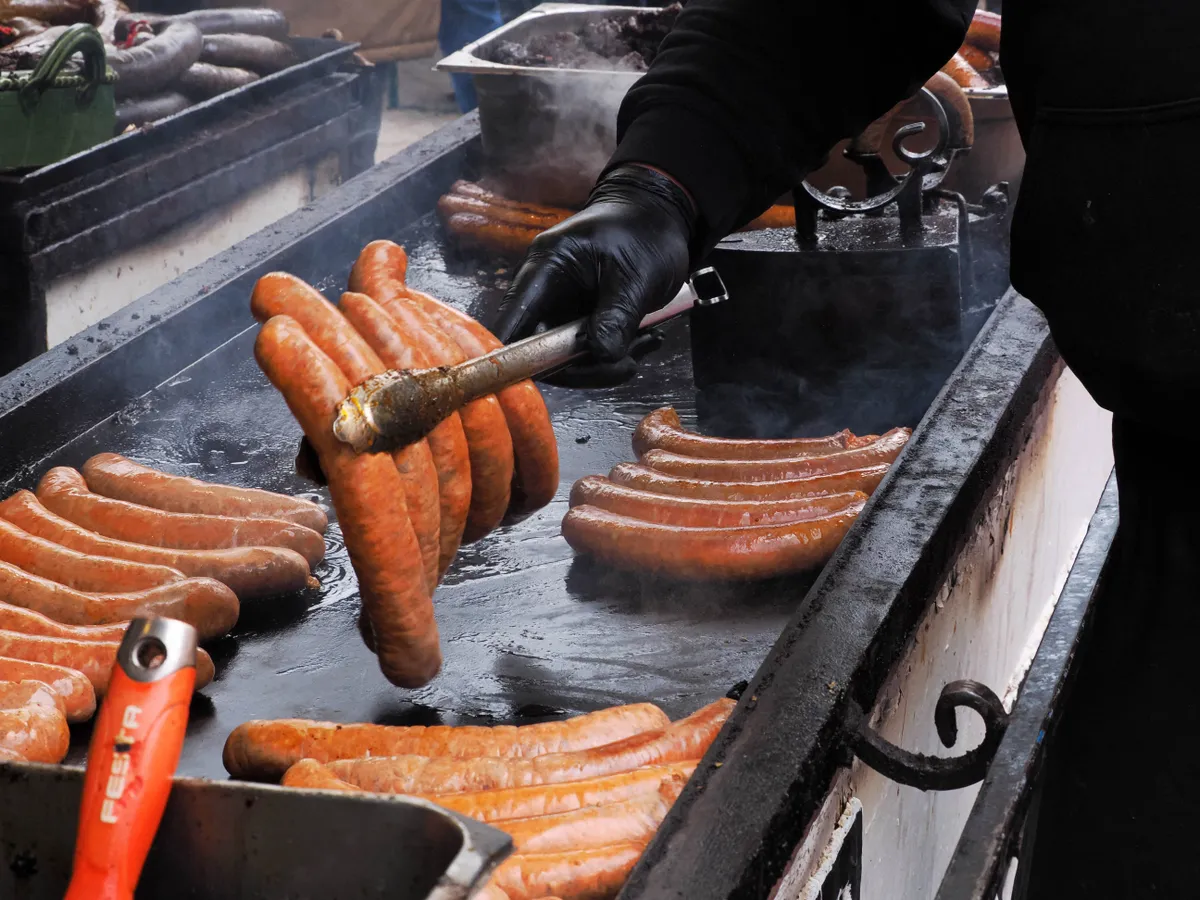 Kolbászok sülnek vaslapon, egy kéz forgatja őket.