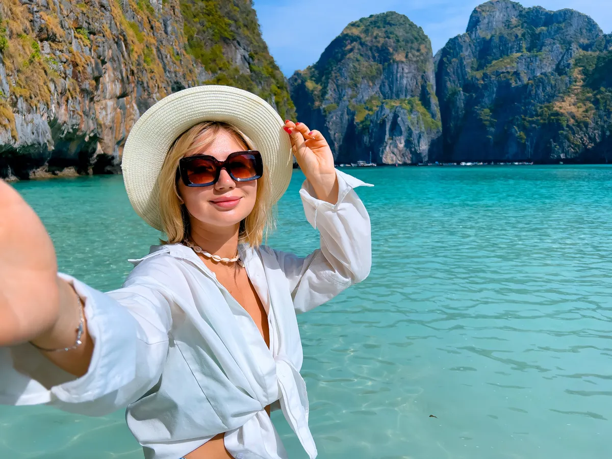 Utazás olcsón: napszemüvegben szelfiző szőke nő egy gyönyörű tengerparton