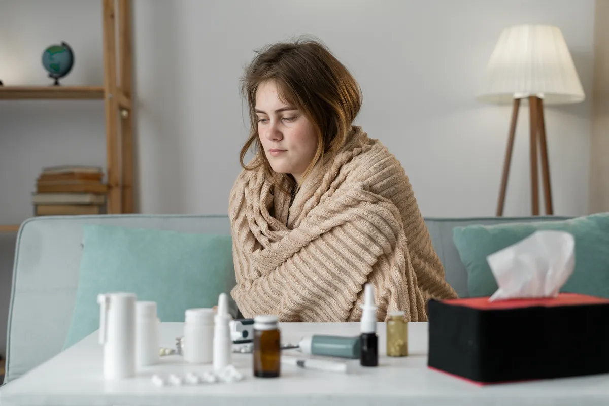 Lázas nő takaróba bugyolálva a kanapén ülve, előtte gyógyszerek