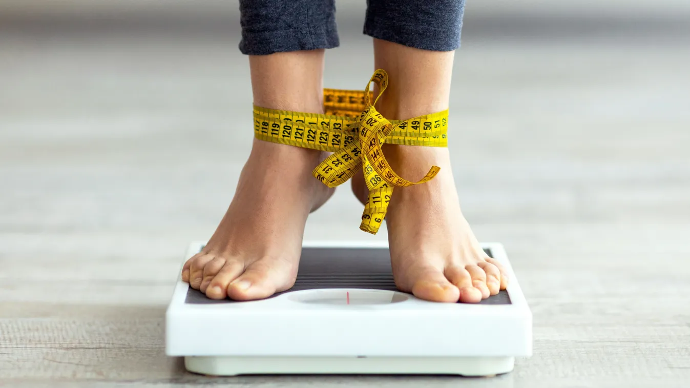 Sikertelen fogyókúra illusztrációja egy mérlegen álló, mérőszalaggal a bokájánál megkötött nővel