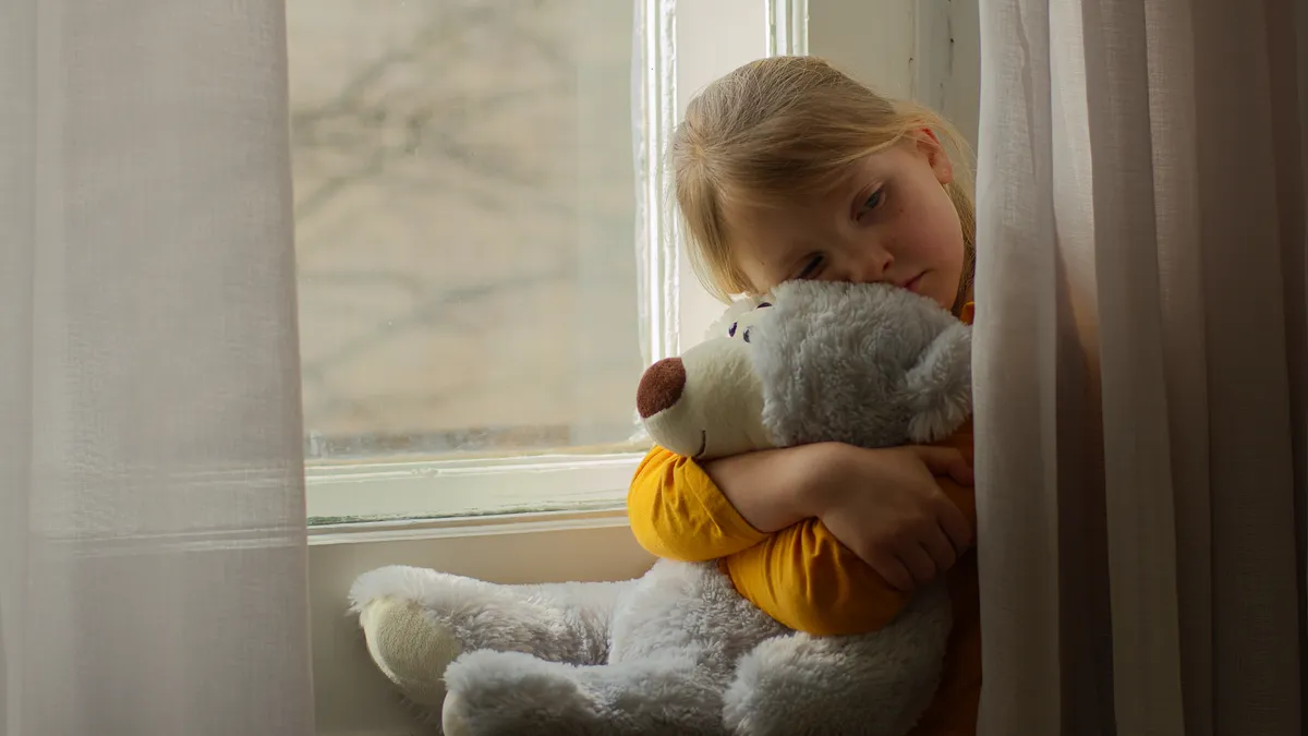 Kedvenc gyerek miatt szomorkodó kislány az ablakban öleli  a plüssmaciját.