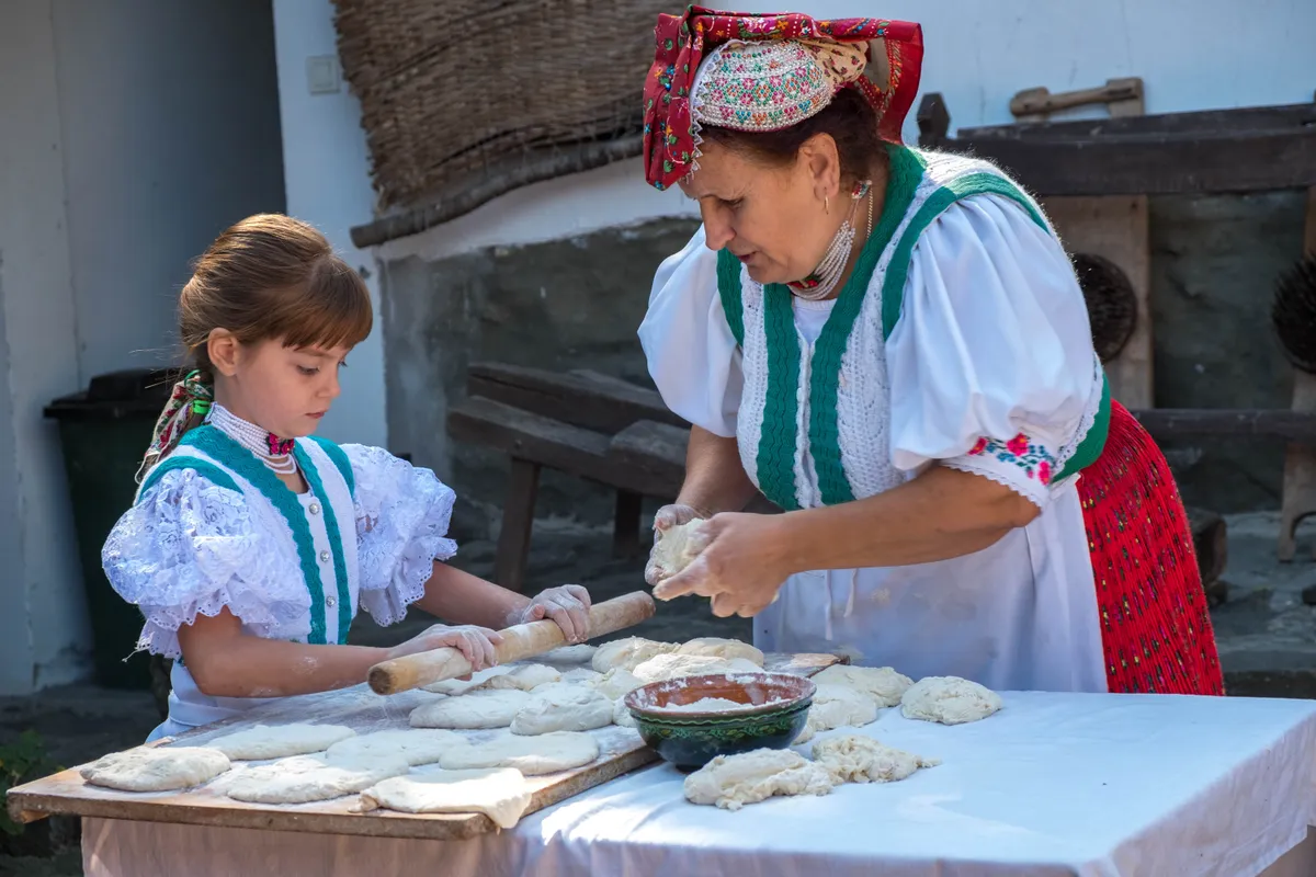Két nő hagyományos magyar népviseletben, tésztagyúrás közben egy kültéri asztalnál, készülve egy műveltségi kvízesthez