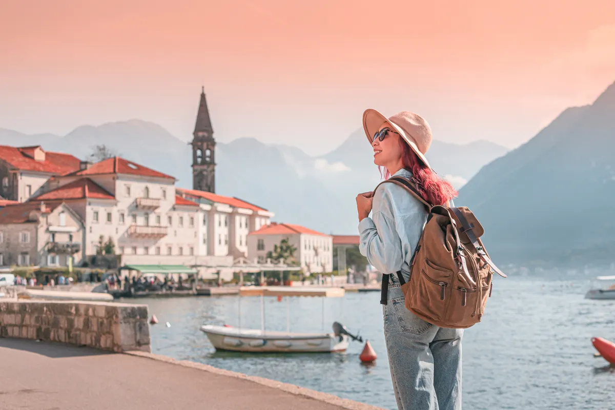 Fiatal hátizsákos nő áll talán montenegrói tengerparti cáros partján, hátérben csónakok, templomtorony.