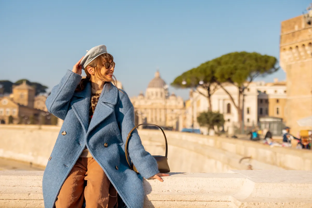 Fiatal nő kép kabátban, svájci sapkában, napszemüvegben ül egy hídn, háttérben római háttérrel.