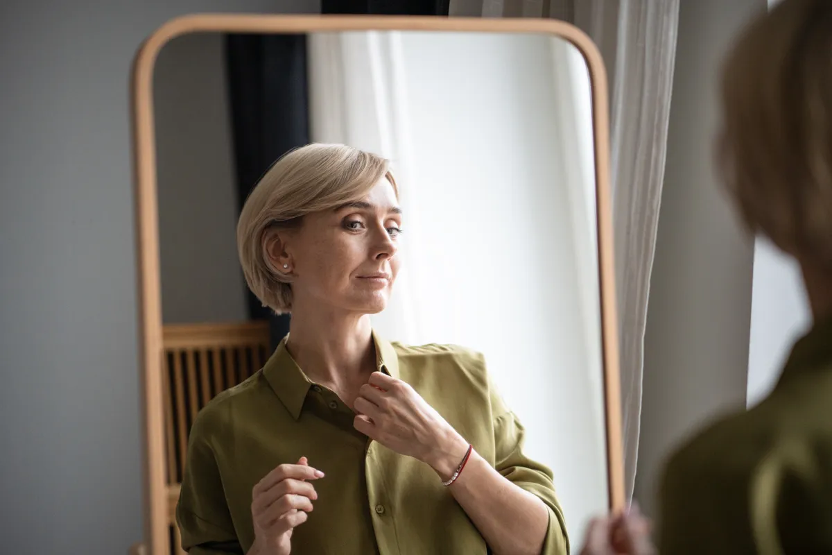 Egy tükörben önmagát néző divatos középkorú szőke nő zöldessárga ingben, aki tudja, melyek a legjobb stílustippek 50 felett