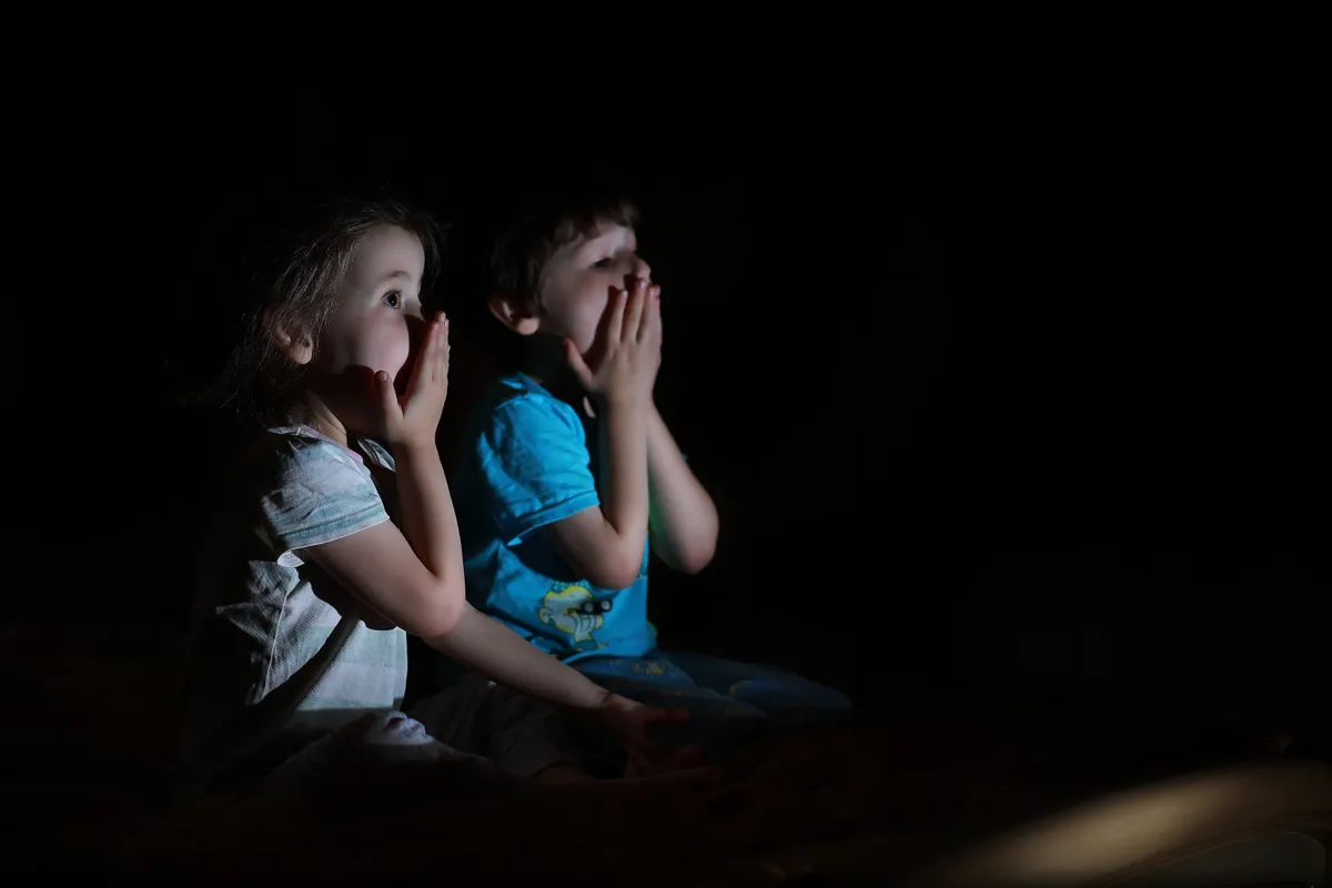 Két kisgyerek tévét néz, a kezüket a szájuk elé tették.