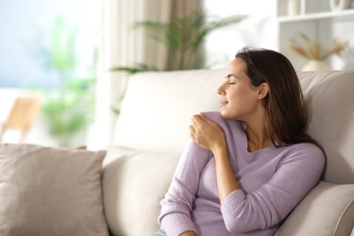 Fiatal nő lilás színű pulóverben a kanapén ülve és azon elmélkedve, hogyan hat rá a tárgyak energiája