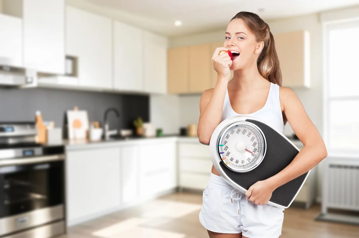 Fogyókúrás tévhitekkel tisztában lévő nő almaevés közben, mérleggel a kezében a konyhában állva