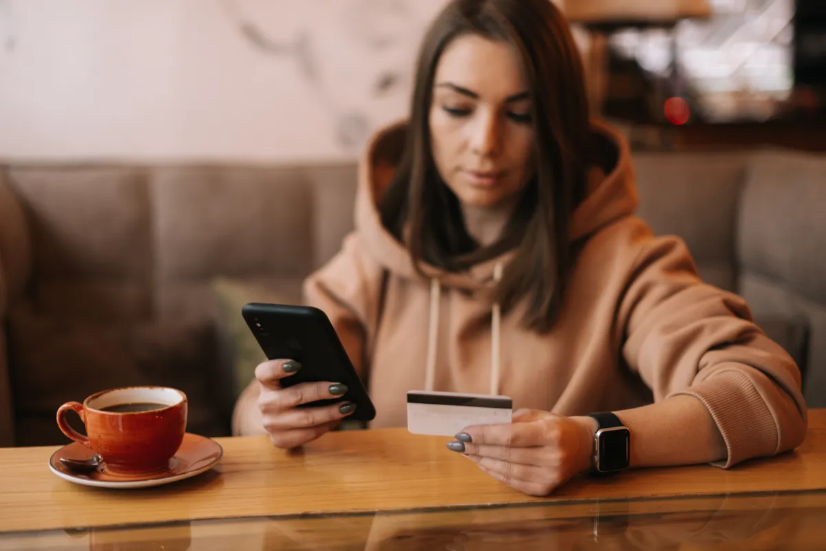 Bankkártyaadatait a mobilbankján ellenőrző fiatal nő egy kávézóban