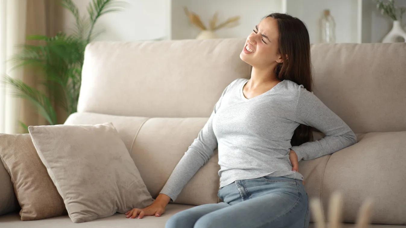 Hátában izomgörcsöt érző nő fájdalmas tekintettel a hátához kapva egy kanapén