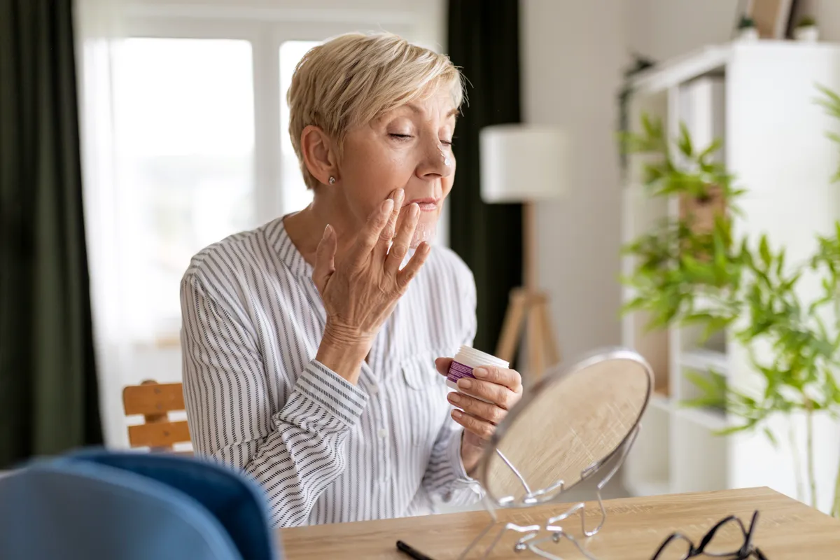 Középkorú nő küzd a menopauza tüneteivel és krémezi az arcát.