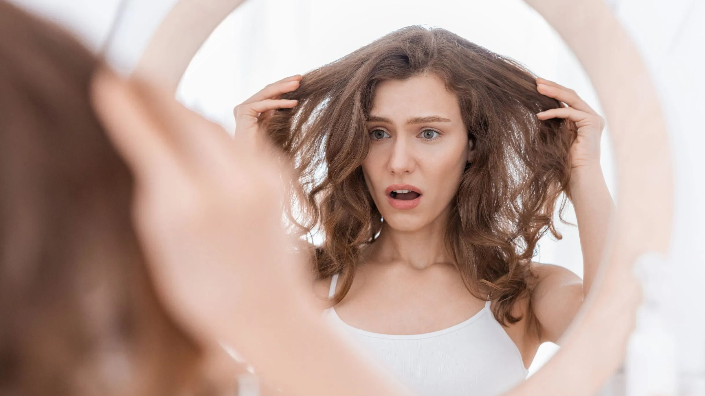 A haját a tükörben aggódva vizsgáló fiatal nő, aki rossz hajápolási rutint követ