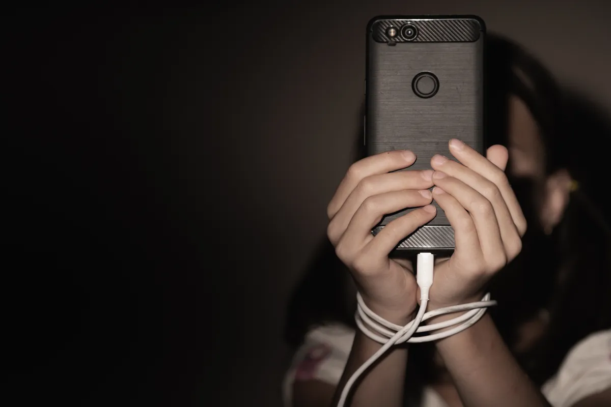 Személyiségtesztben szereplő kép egy fiatal lányról, akinek a kezéhez van kötözve egy okostelefon annak töltőzsinórjával a telefonfüggőség szimbolizálásaként