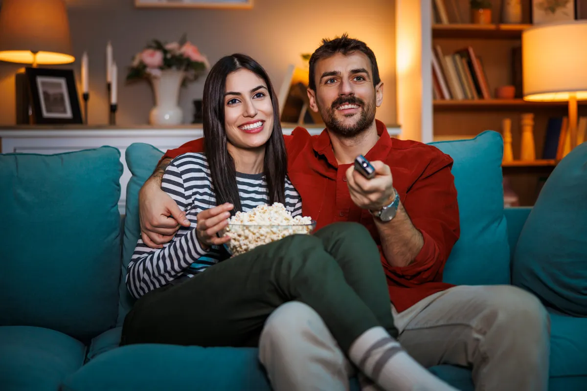 Filmes kvízben szereplő kép egy kanapén ülő boldog fiatal párról filmezés közben egy tál pattogatott kukoricával