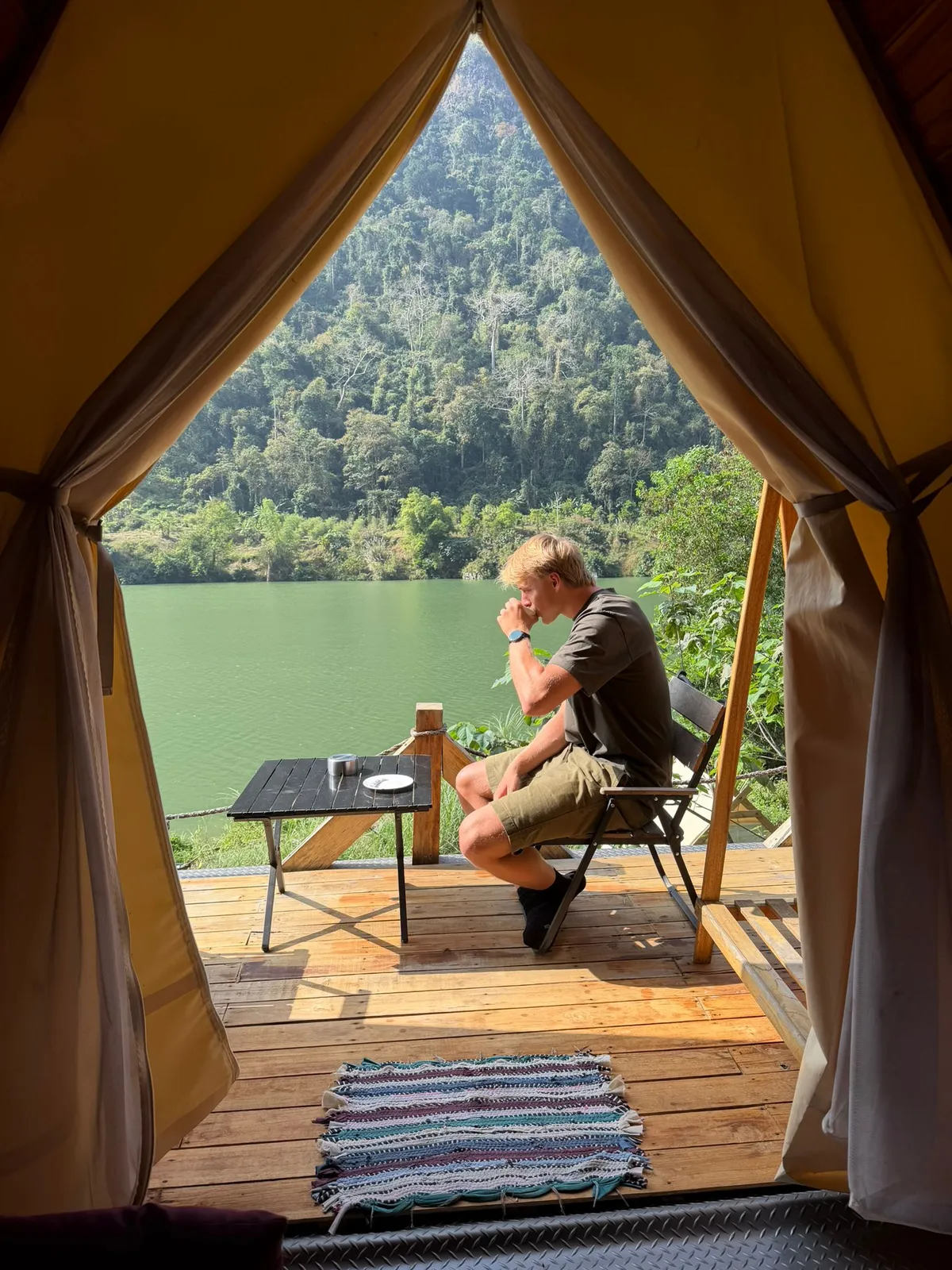 Gergő egy sátor előtt ül és iszik, háttérben tenger és délkele-ázsiai növényzet.