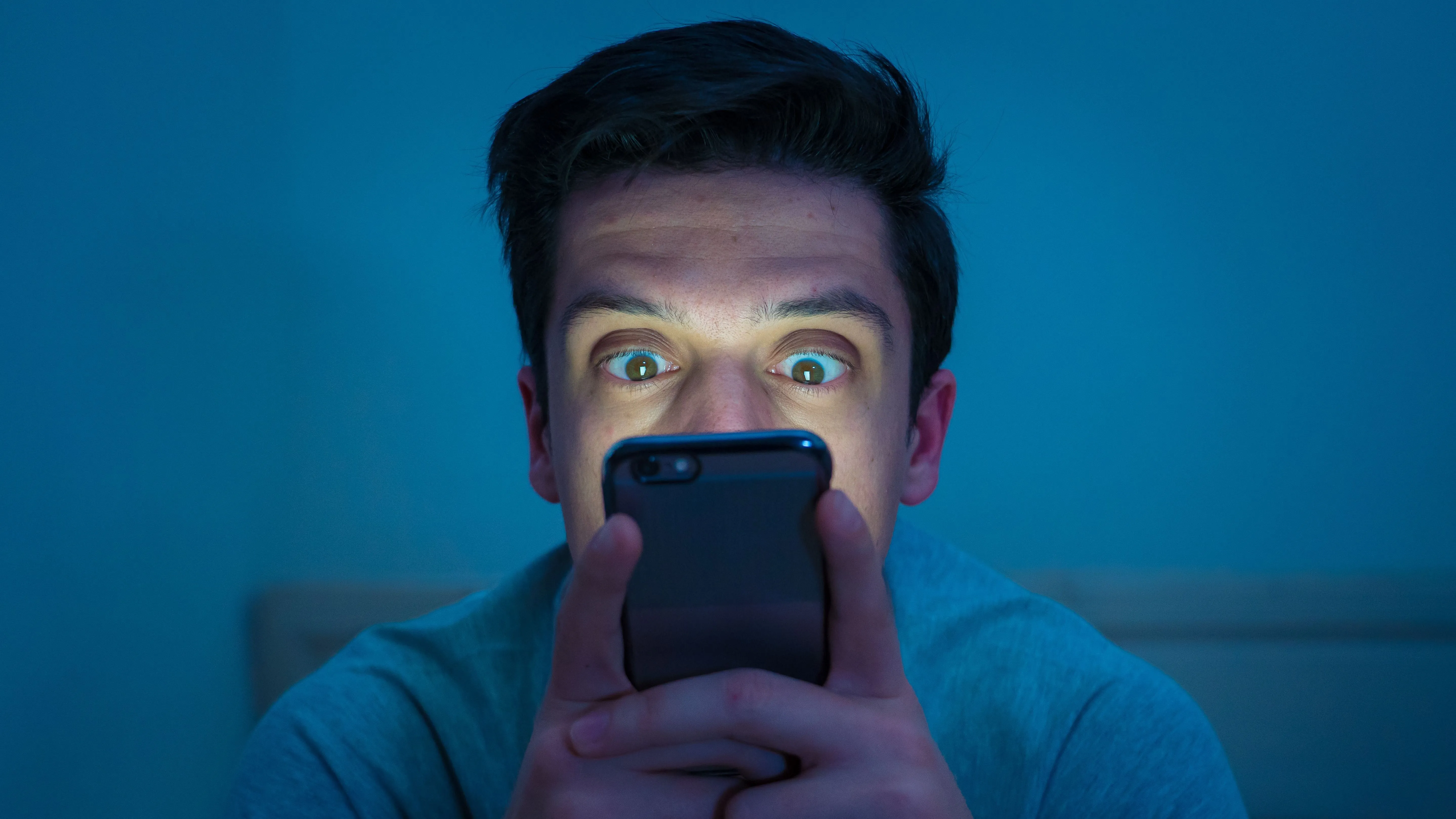 Az okostelefonján telefonfüggőségi személyiségtesztet töltő telefonfüggő fiatal férfi, akinek az arcát megvilágítja a készülék képernyőjének fénye